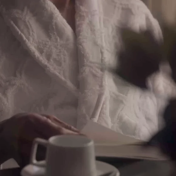 A man in a white bathrobe and glasses reads a paper while sitting at a table with a white coffee cup.