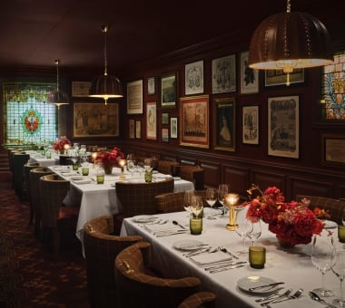 An elegantly appointed private dining room featuring long tables set with crisp white tablecloths, glowing gold cordless lamps, green glassware, and vibrant red floral centerpieces