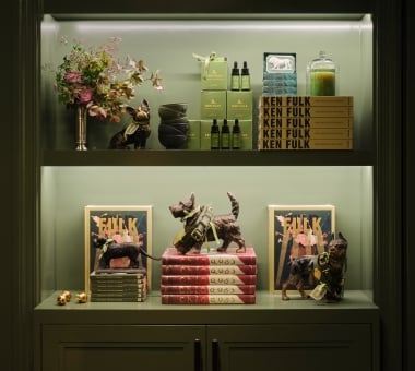 A carefully arranged, multi-shelf display cabinet in a muted olive green color features structured stacks of books, small dog figurines, and luxury lifestyle products