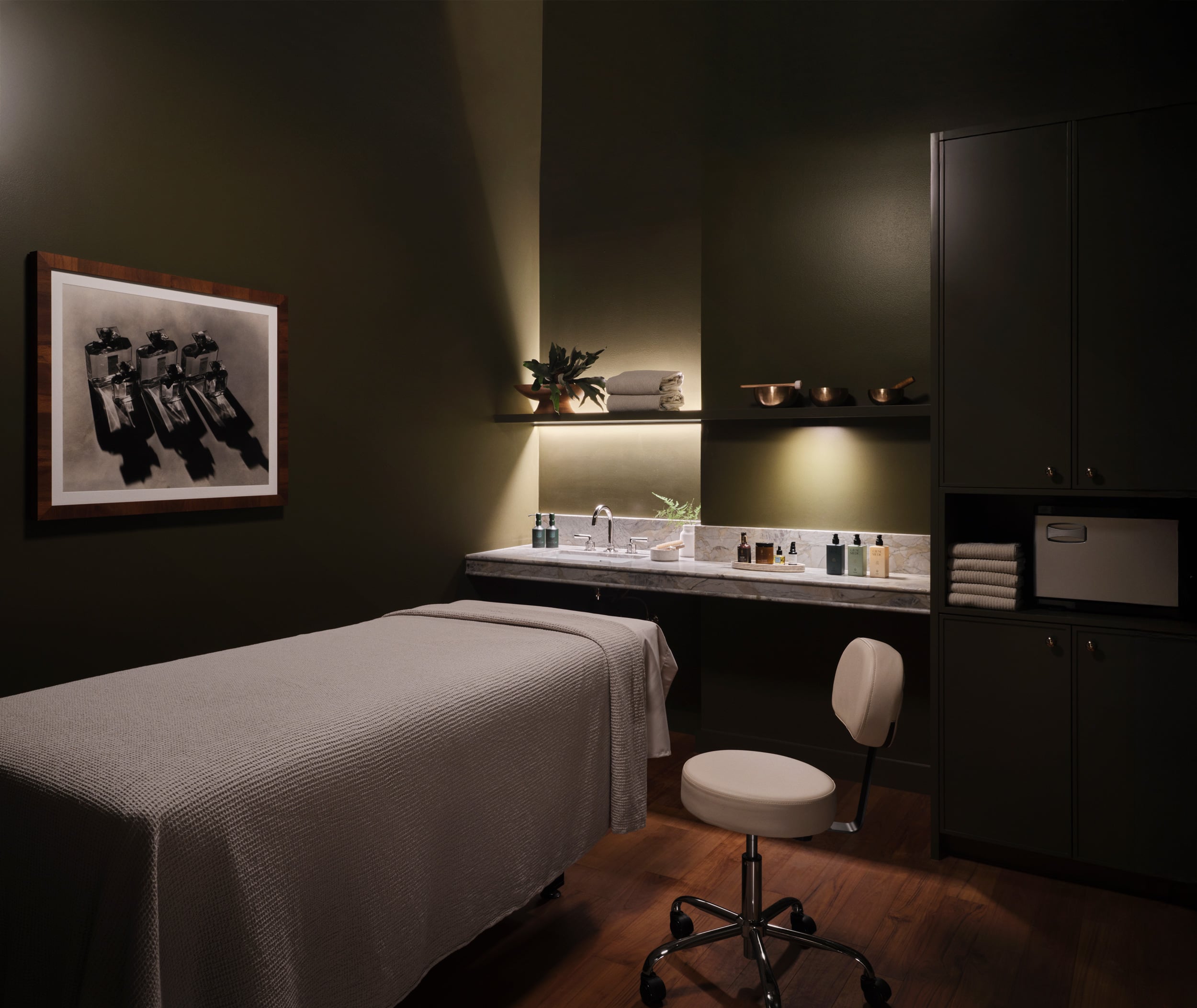 A well-lit spa treatment room with dark green walls, a draped massage table, a rolling rolling stool