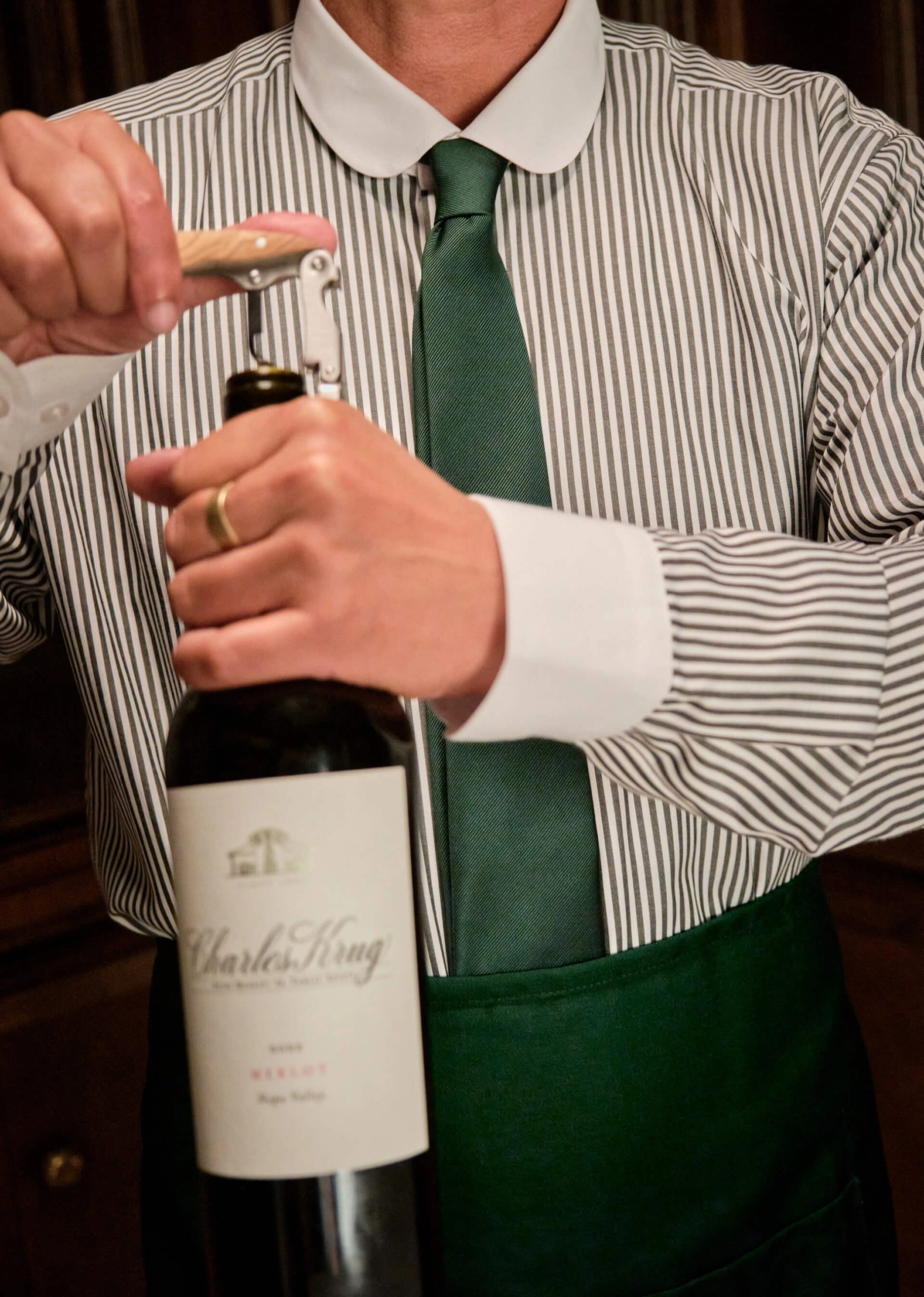 A server in a striped shirt and green tie opening a bottle of Charles Krug wine