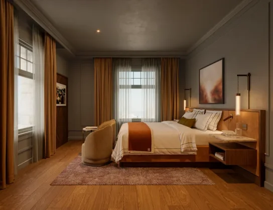 A hotel room with slate gray walls accented by rich ochre textiles, features elegant details such as wall wainscotting, marble-topped night stands, pendant lighting, and wide-plank wood floors.