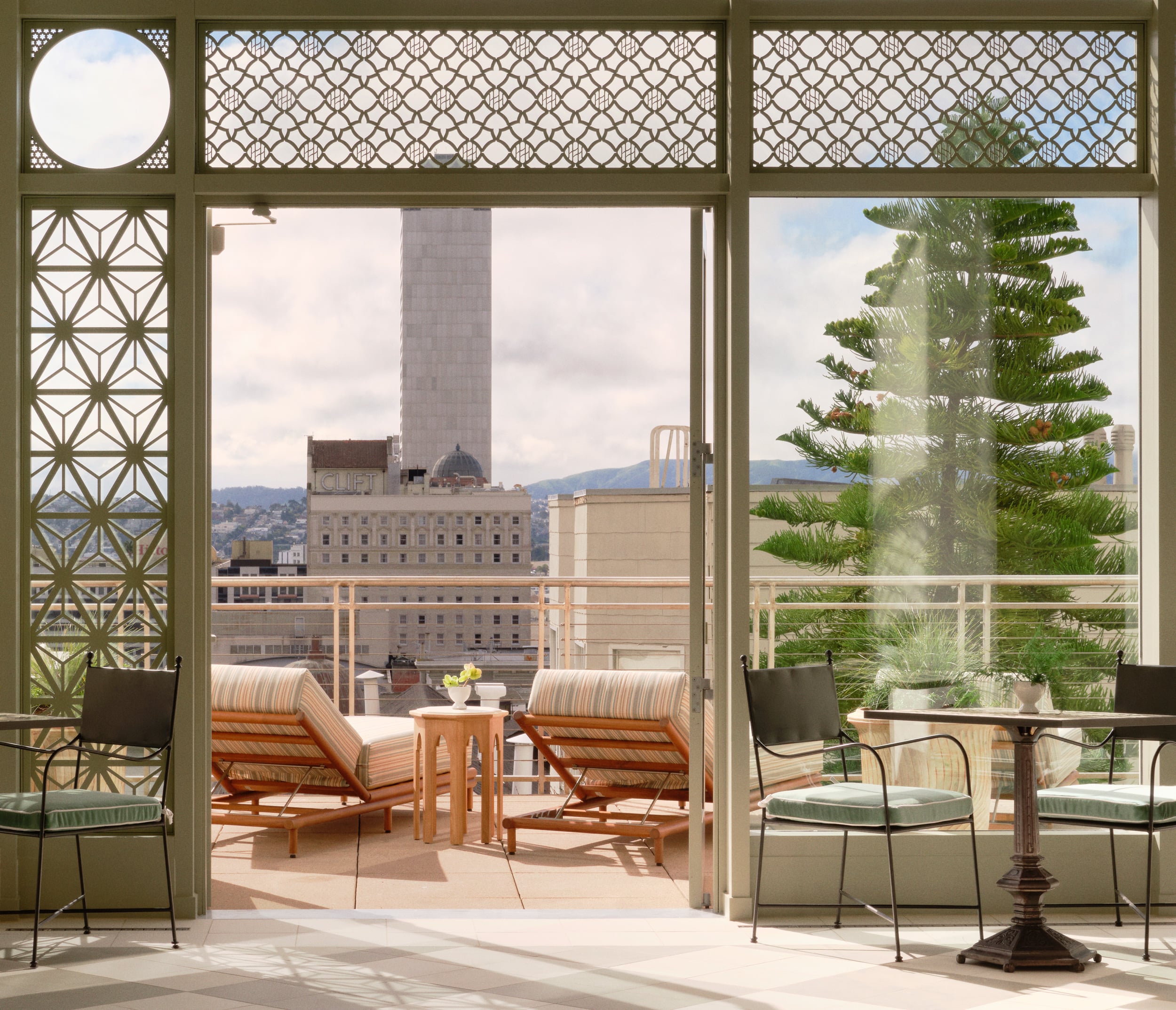 A sunlit indoor terrace with olive green geometric lattice screen panels opens up to an outdoor balcony overlooking a city skyline