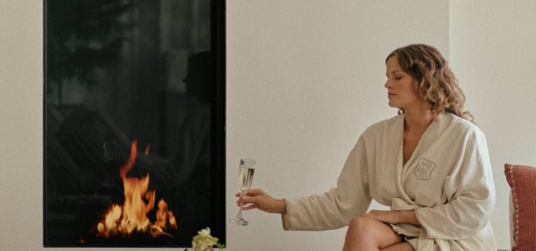 A woman in a robe sitting by the fireplace with a glass of champagne and cheese board