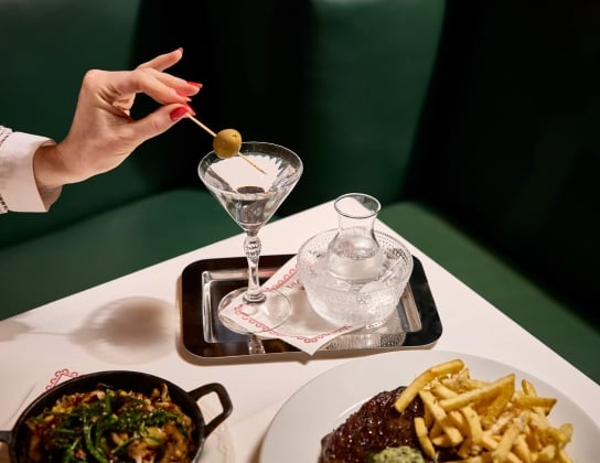 Hand placing Martini Olive Garnish in drink with plates of food surrounding