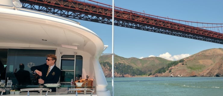 A yacht sailing under the Golden Gate bridge as a bartender opens a bottle of wine.
