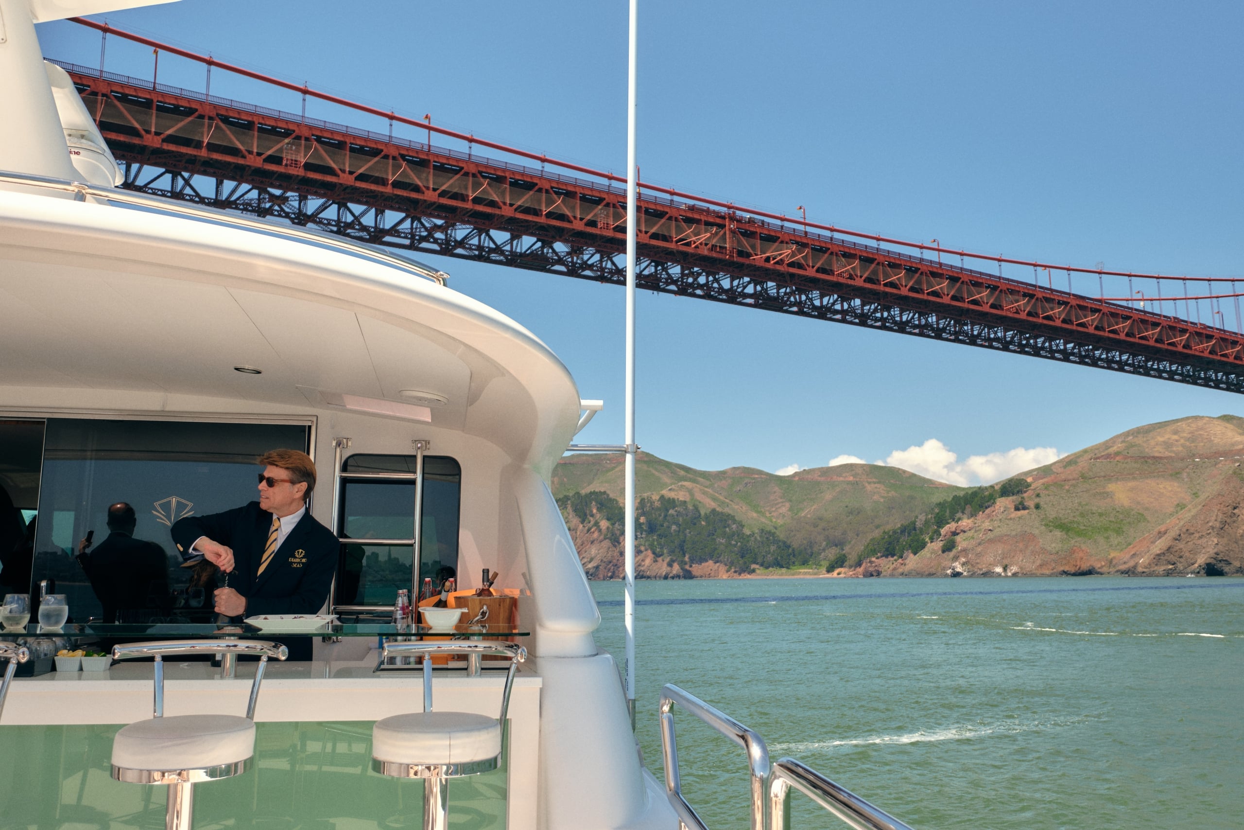 A yacht sailing under the Golden Gate bridge as a bartender opens a bottle of wine.