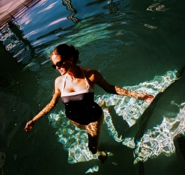 high-angle shot captures a woman wading in a swimming pool, with the water reflecting dark green tones and rippling sunlight