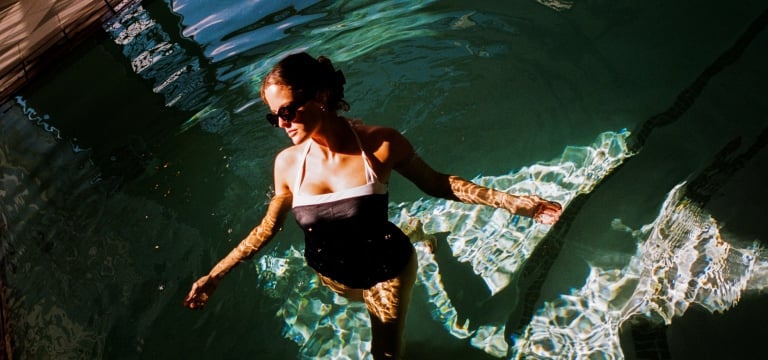 high-angle shot captures a woman wading in a swimming pool, with the water reflecting dark green tones and rippling sunlight