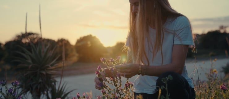 A young person with long hair stands outdoors at sunset, gently holding a sprig of flowers