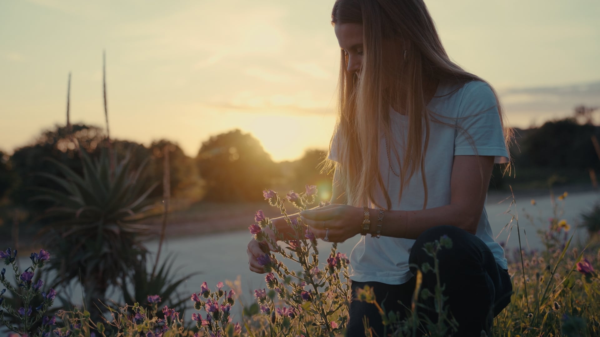 A young person with long hair stands outdoors at sunset, gently holding a sprig of flowers